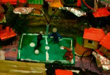 Nesta foto em close, vê-se fração de maquete na qual se reconhece ambiente de uma comunidade ou favela. Rodeada por pequenas casas e prédios coloridos, há uma quadra de esportes. Nota-se ainda que as moradias foram erguidas sem planejamento.