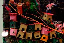 Esta foto em close revela fração de maquete na qual se reconhece ambiente de uma comunidade ou favela. Nota-se que as pequenas casas coloridas foram erguidas sem planejamento, ocupando os contornos do monte. Percebem-se ainda alguns fios, também desordenados, representando as ligações caseiras que levam energia elétrica às moradias.