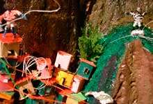 Parte de um tipo de maquete é mostrada nesta foto. Nesta foto, veem-se recriações de pequenas casas coloridas, erguidas sem planejamento e encostadas em rocha de um morro.