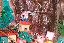 Parte de um tipo de maquete é mostrada nesta foto. Veem-se recriações de pequenas casas coloridas, erguidas sem planejamento e encostadas em rocha de um morro. Mexendo nos vários fios amontoados, vê-se uma pessoa no alto, próxima à rocha.