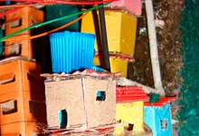 Esta foto em close revela fração de maquete na qual se reconhece ambiente de uma comunidade ou favela. Nota-se que as pequenas casas coloridas foram erguidas sem planejamento, ocupando os contornos do monte. Percebem-se também alguns fios, também desordenados, representando as ligações caseiras que levam energia elétrica às moradias.